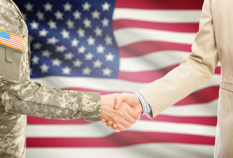 U.S. military person shaking hands with business person in front of United States flag.