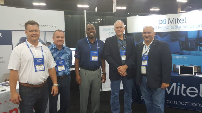 A grouping of four male Frontier and Mitel employees standing in front of their company's convention tables.