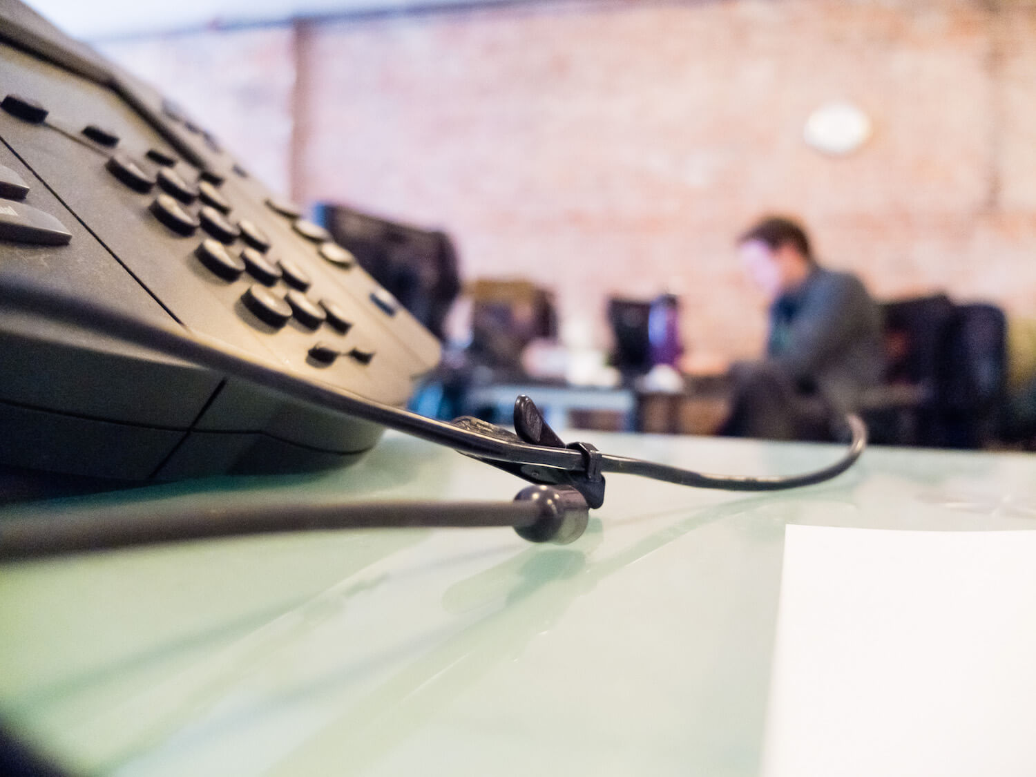 Business VoIP telephone system with cord leading to blurred image of man sitting at desk in background.